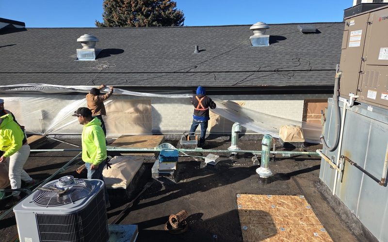 Roofing crew in safety gear masking a building's exterior brick wall with plastic sheeting to prep for work, on a commercial rooftop with HVAC units, vent pipes, and a shingled roof section under a clear blue sky.