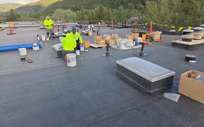 Roofing crew in safety gear masking a building's exterior brick wall with plastic sheeting to prep for work, on a commercial rooftop with HVAC units, vent pipes, and a shingled roof section under a clear blue sky.