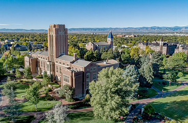 Aerial view of a university campus building with a clock tower, surrounded by trees and mountains in the background