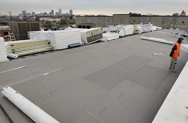 Worker walking a flat roof membrane installation with insulation rolls staged and a city skyline in view