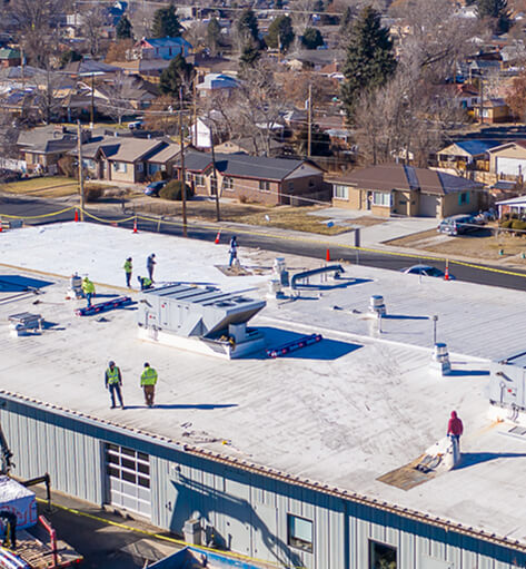 Aerial view of a roofing crew replacing a white membrane roof on a commercial building in a residential neighborhood