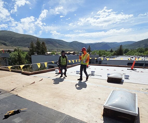 Roofing crew in safety vests installing single-ply membrane on a commercial roof with mountains in the background