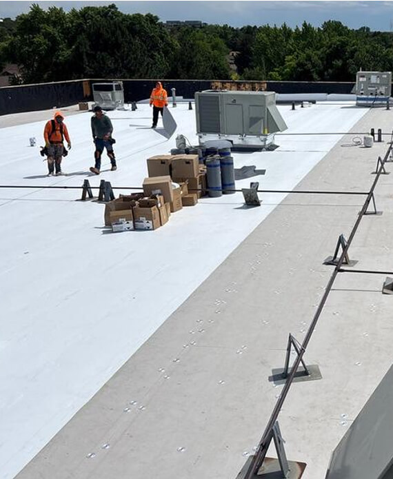 Roofing crew installing a white TPO membrane roof with materials staged and a warning-line system along the perimeter