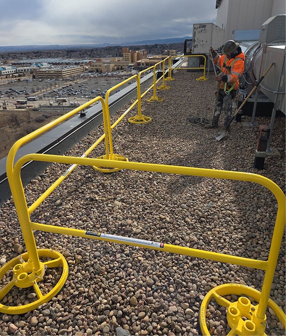 Worker in high-visibility vest performing rooftop maintenance behind a yellow freestanding safety guardrail