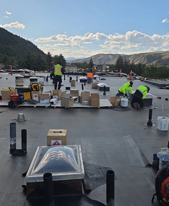 Roofing crew installing EPDM membrane and detailing penetrations on a commercial rooftop with mountain views